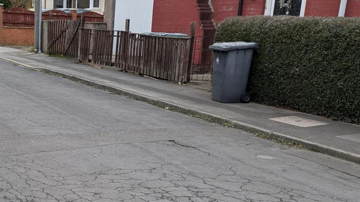 A grey bin outside a house, which has a hedge and red bricks