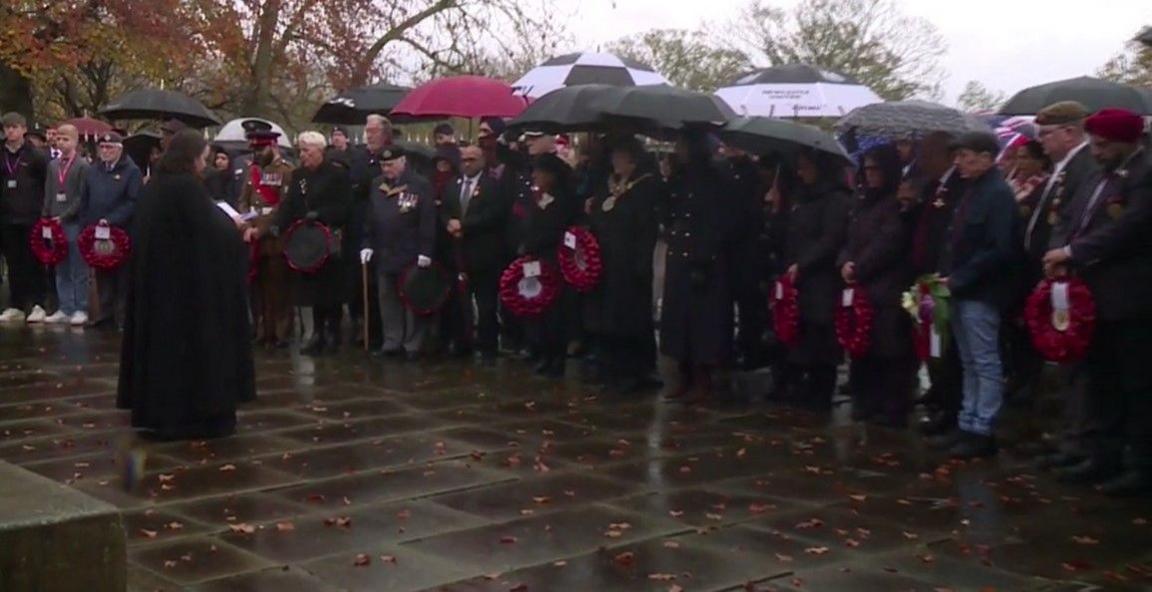 A large group of people gathered on a paved area outside. Many are holding black umbrellas, while others have red poppy wreaths in their hands. Several individuals wear formal attire, including dark coats and military uniforms with medals. 
