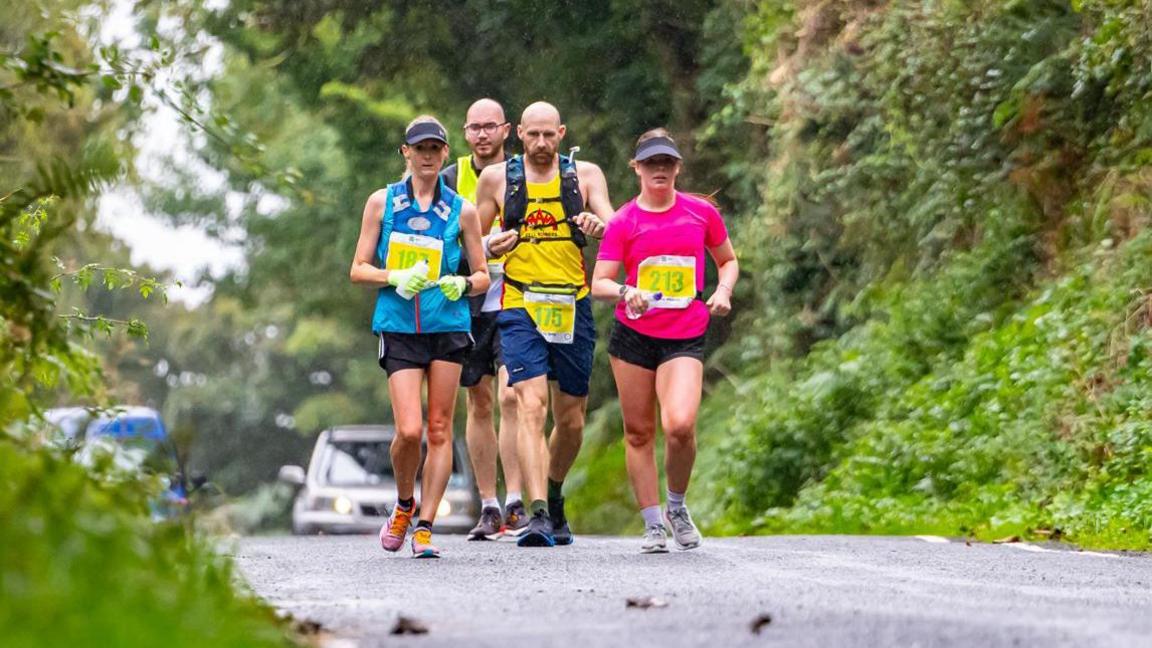 Four walkers racing through a green lane. Two women, one wearing a pink T-shirt, another wearing a blue vest, are leading the pack with two men following. Two cars can be seen parked up in the distance behind them.