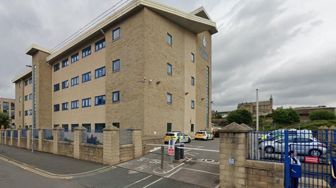 A four-storey modern beige brick building. Police cars are parked in the car park outside it, which is barriered off.