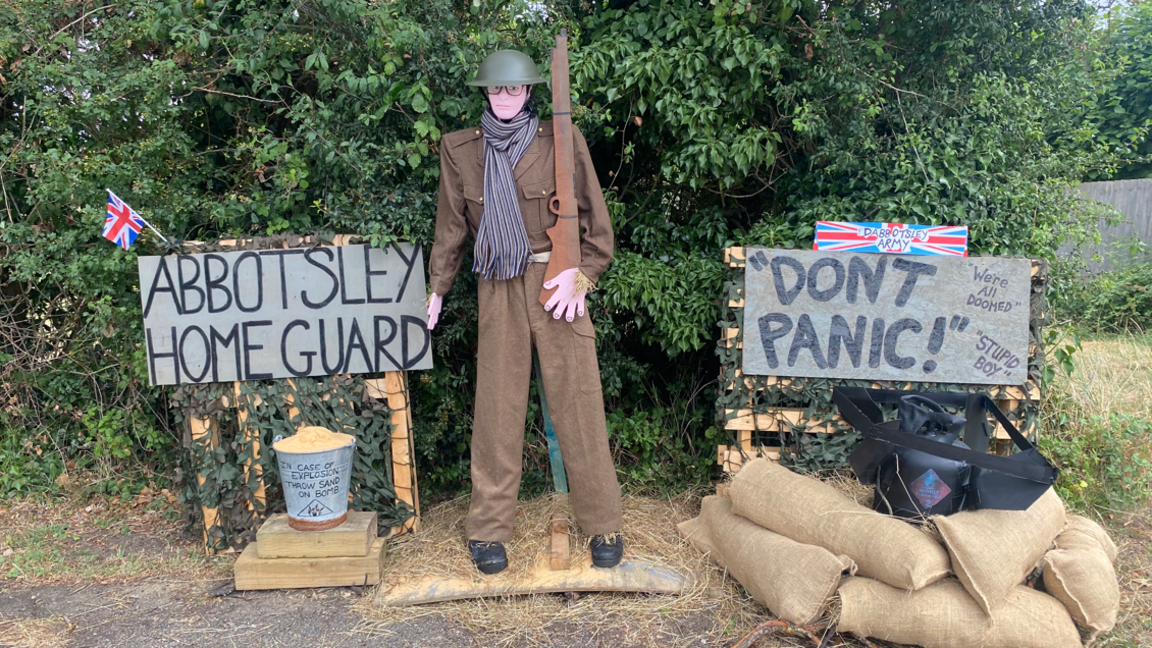 A life-size scarecrow of a soldier dressed in a brown uniform, green helmet, with glasses. It is holding a gun and has a scarf around its neck. On the ground are four sandbags and two signs. One reads 'Abbotsley home guard' and the other says 'Don't panic', 'Stupid boy' and 'We're all doomed'. There is also a bucket on the ground which says 'in case of explosion, throw some sand on bomb'. 
