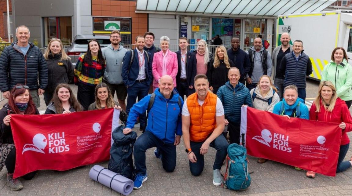 More than 20 people posing for the camera outside a building. Mike Weaver (centre right) has one knee on the ground and is wearing an orange top. Two identical red and white banners, including the words Kili for kids, are being held by other people. 