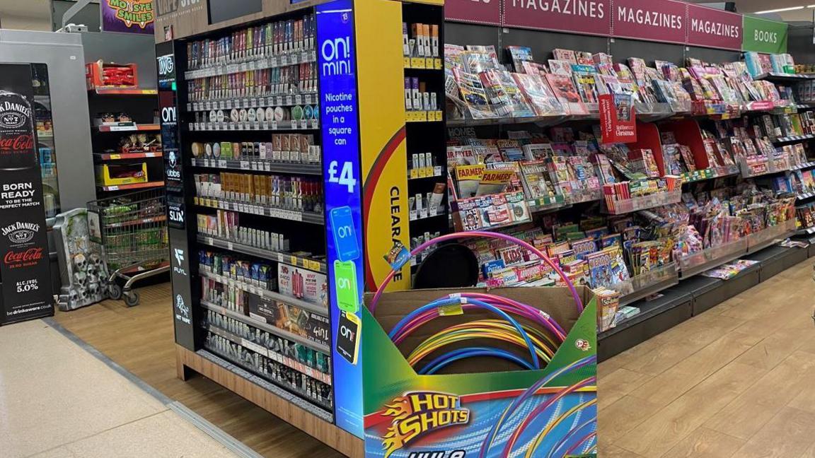 A display on the end of an aisle showcasing colourful boxes of vapes. On the side there is a section of “clearance” vapes, with children's hula hoops positioned right in front of them. Further down the aisle there are magazine displays. 