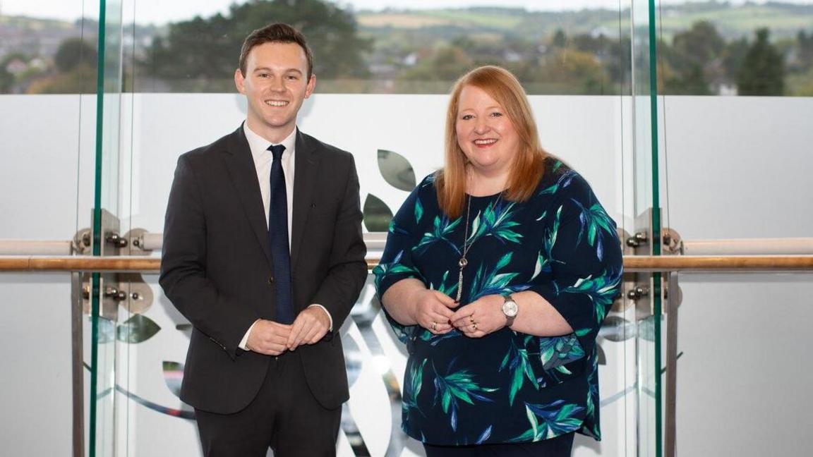 Eoin Tennyson and Naomi Long - Eoin wearing a black suit, white collared shirt and blue tie stands with his hands held in front of his body. On his right is a Naomi who is shorter, ginger haired and wearing a blue and green floral patterned shirt. They are standing in front of frosted glass windows and smiling at the camera.