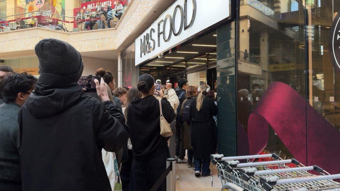 A crowd of people taking photographs with their mobile phones outside a Marks and Spencer store. In the foreground can be seen shopping trolleys.
