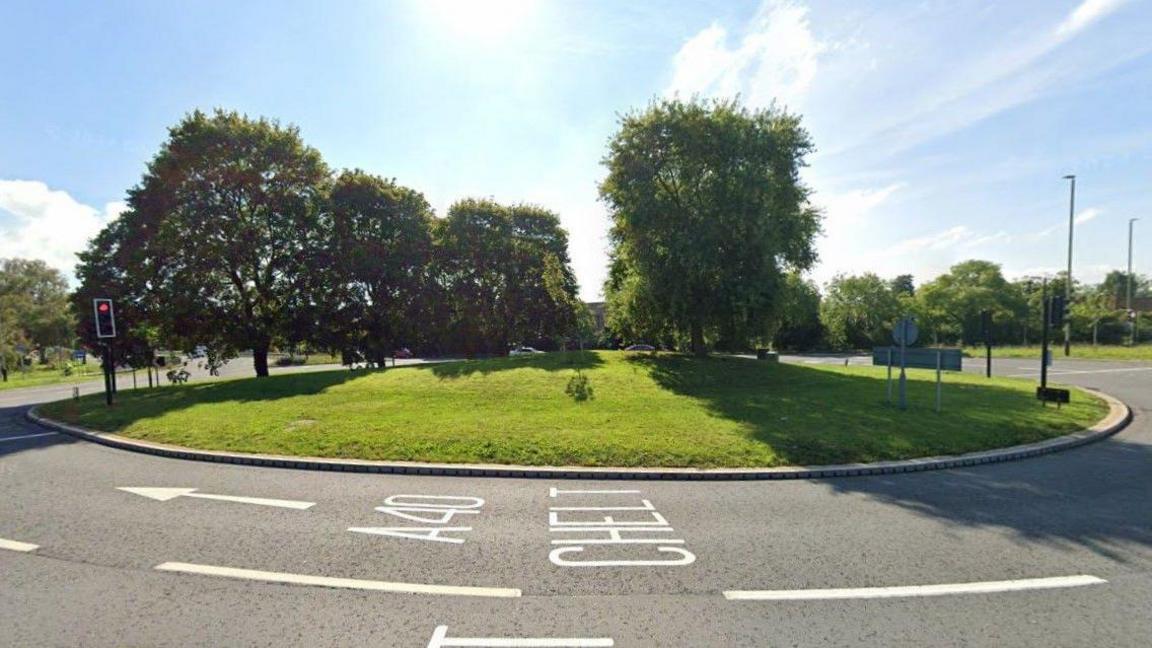 A screenshot from Google street view showing the A40 Benhall Roundabout in Cheltenham. It is a sunny day and the roundabout mound is covered in grass and several trees. There are no cars on the road at the time the picture was taken.