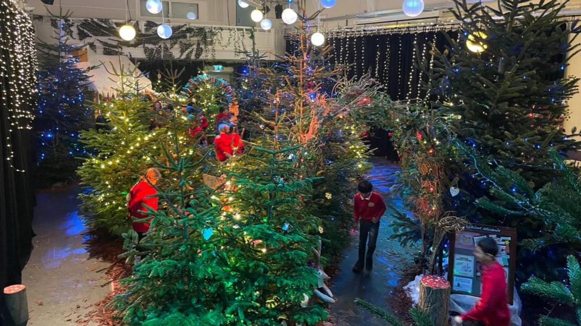 A former church hall is decorated with Christmas trees and fairy lights. Primary school children run around the theatre set dressed in their red school uniforms. 