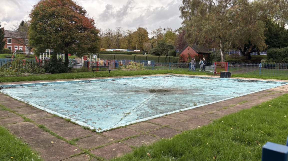 A shallow paddling pool that has been completely drained and is now in disrepair with dirt of vegetation growing on it.