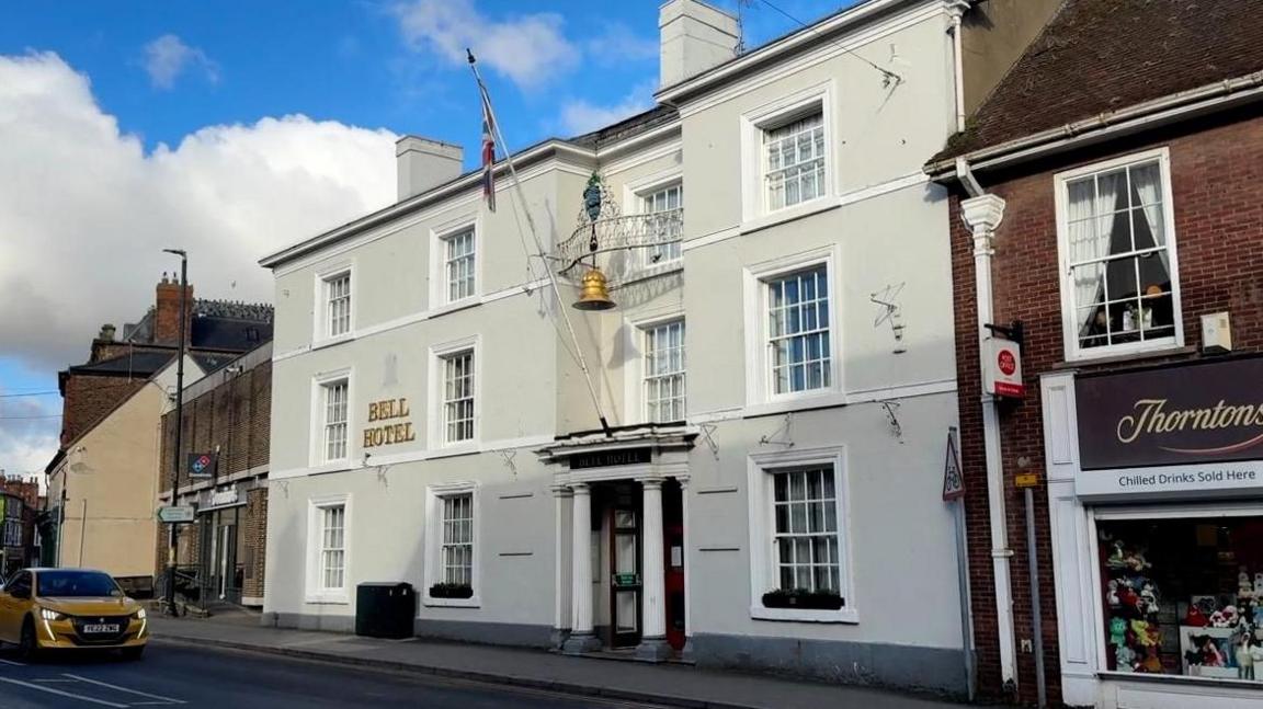 The exterior of the Bell Hotel in Driffield. The Grade II listed building has been painted grey-white and has large sash windows. A golden bell hangs above the main entrance which also has a pole flying the union flag.