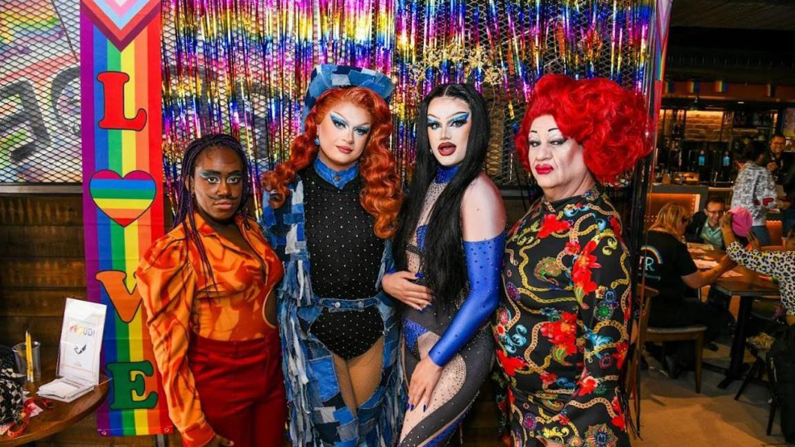 Drag queens and kings posing in front of a rainbow banner containing the word "love"