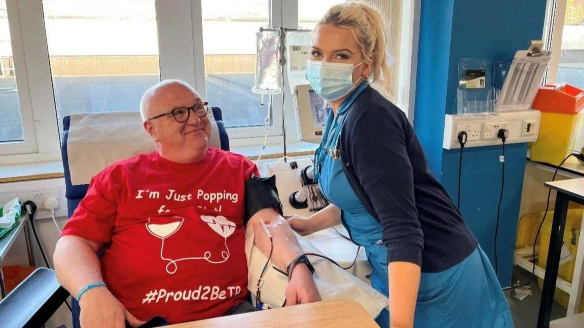 A blood donor lying down with a tube in his arm. He is wearing a red t-shirt saying he is 'proud to be a donor'. Somer is standing next to him wearing a mask and checking he is well. 