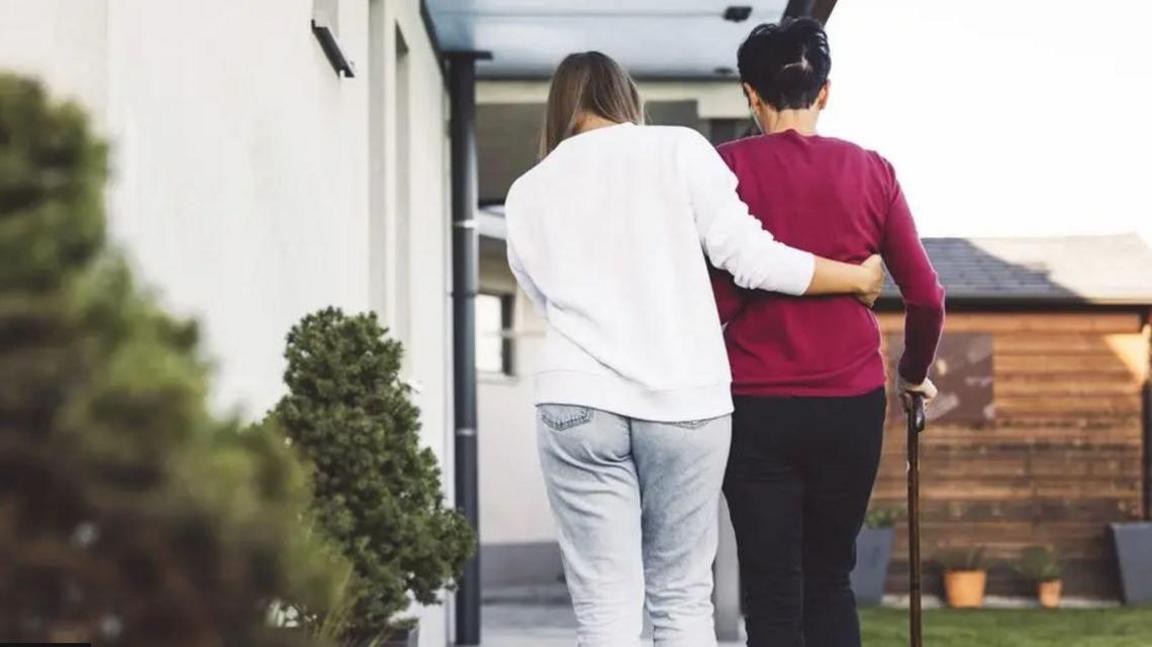 A younger woman dressed in white from behind with her arm around an elder woman in a red top and black trousers using a walking stick. The younger woman seems to be helping the older woman up a pathway to a house