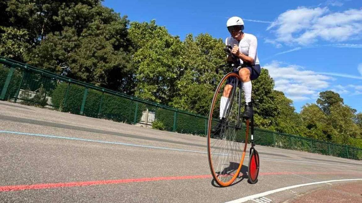Richard Thoday on an orange framed penny farthing at Reading Velodrome. The picture has been taken from ground level looking up to Thoday who is wearing lycra and a white helmet.