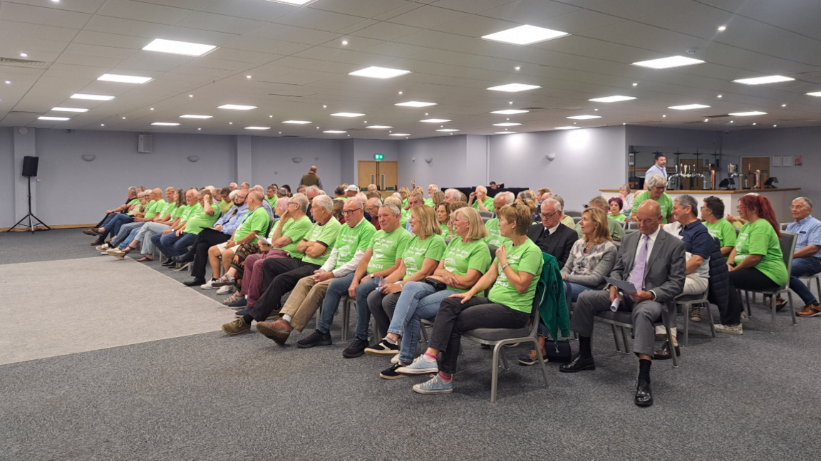 A group of people are sitting in a grey carpeted room with plain walls and a bar at the back. They are sitting on chairs and all face the same direction, to the left of the camera. Many of the people are wearing matching green tops, they are with the Staunch campaign.