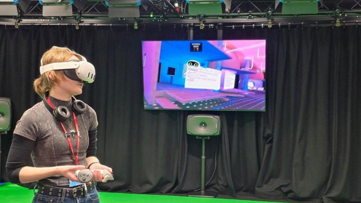 A girl with short ginger hair wears a white VR headset and black headphones around her neck. She holds a white controller in her hands. Behind her is a TV screen showing a CGI image.