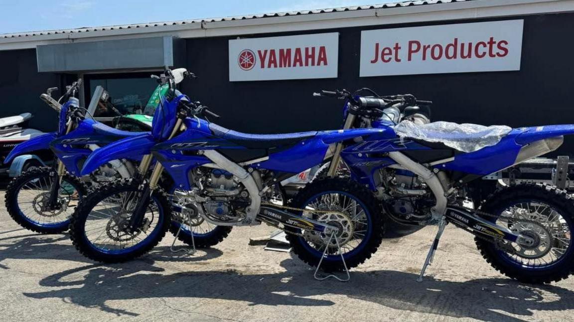 Three blue scrambler bikes are sitting outside the front of a business. The bikes are stood up on grey concrete. The business in the background is a black shed with two large red and white signing reading 'Yamaha' and 'Jet Products' of its front.