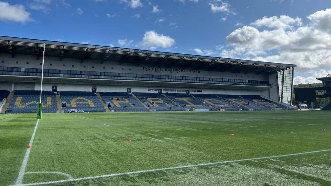 One stand in the background with the word Warriors visible via the yellow seats among the blue seats. The pitch is in the foreground.