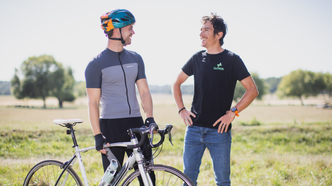 A person is stood on the left wearing a blue and yellow helmet looking at someone to the right of the picture. They are holding a silver bike and wearing a grey zip up sports top and black trousers. Geraint Thomas is on the right looking towards the person on the left smiling. He has short brown hair and a short moustache and beard, and is wearing a black T-shirt with blue jeans. There are trees and a field in the background.