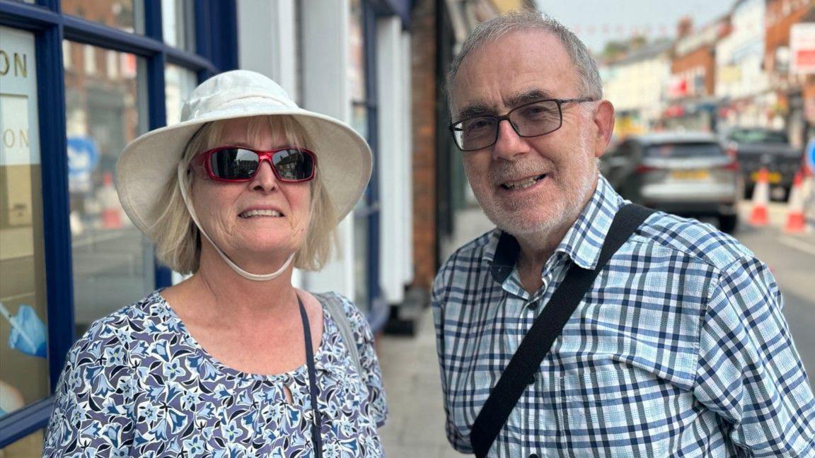 A late middle-aged white couple stand side by side in a high street. She wears a wide-brimmed hat, red sunglasses and a floral top. He is in glasses and a blue checked shirt.