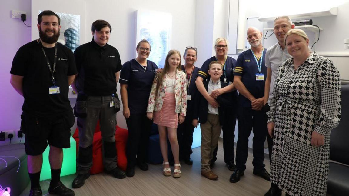 Eight adults and two children stand facing the camera in a sensory room at a hospital 