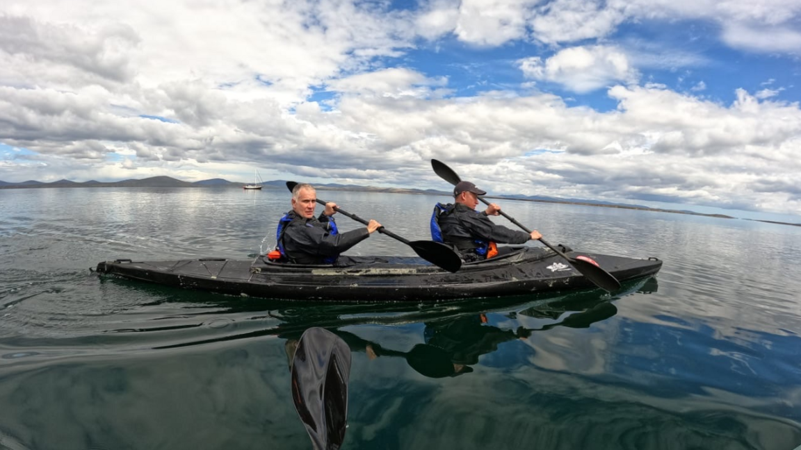 Image shows two men in a kayak on glass like water. They are paddling under a cloud filled blue sky.