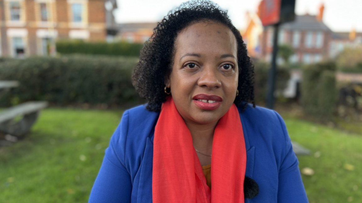 Keisha Adair Swaby, who is wearing a blue blazer and a red scarf, smiles while standing on a lawn outside.