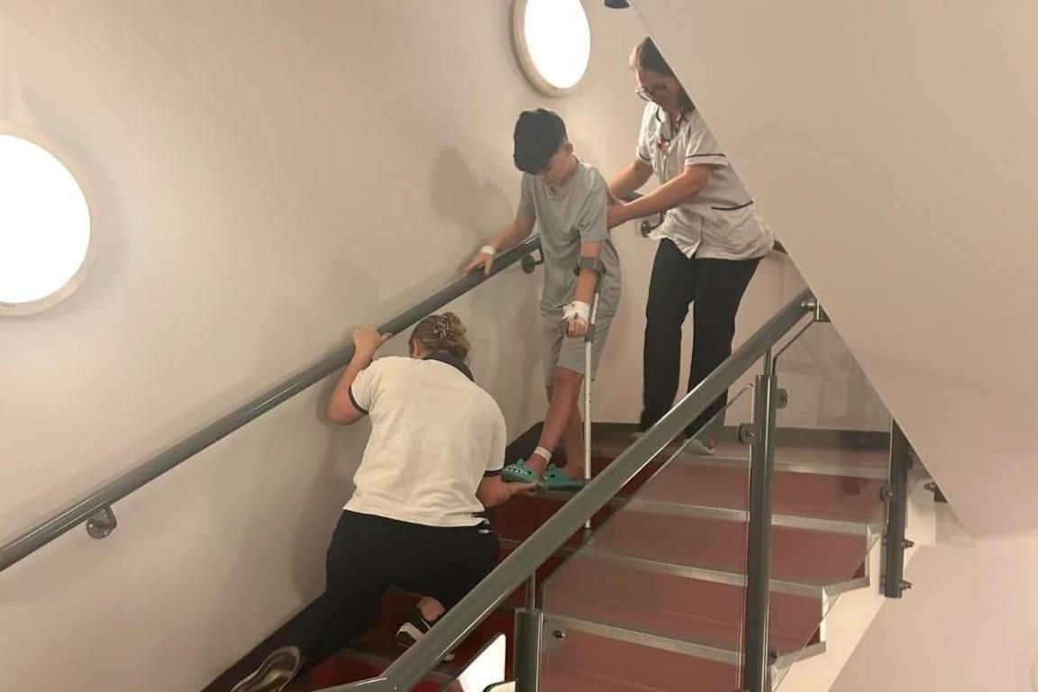 Josh is descending hospital stairs. A nurse is holding him by his back, another is holding his outstretched foot in her hand. 