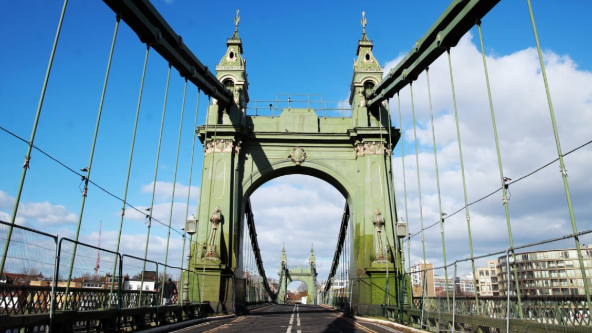 A view of the road and green arches and joists of the bridge on Hammersmith Bridge in 2020, with metal barriers either side