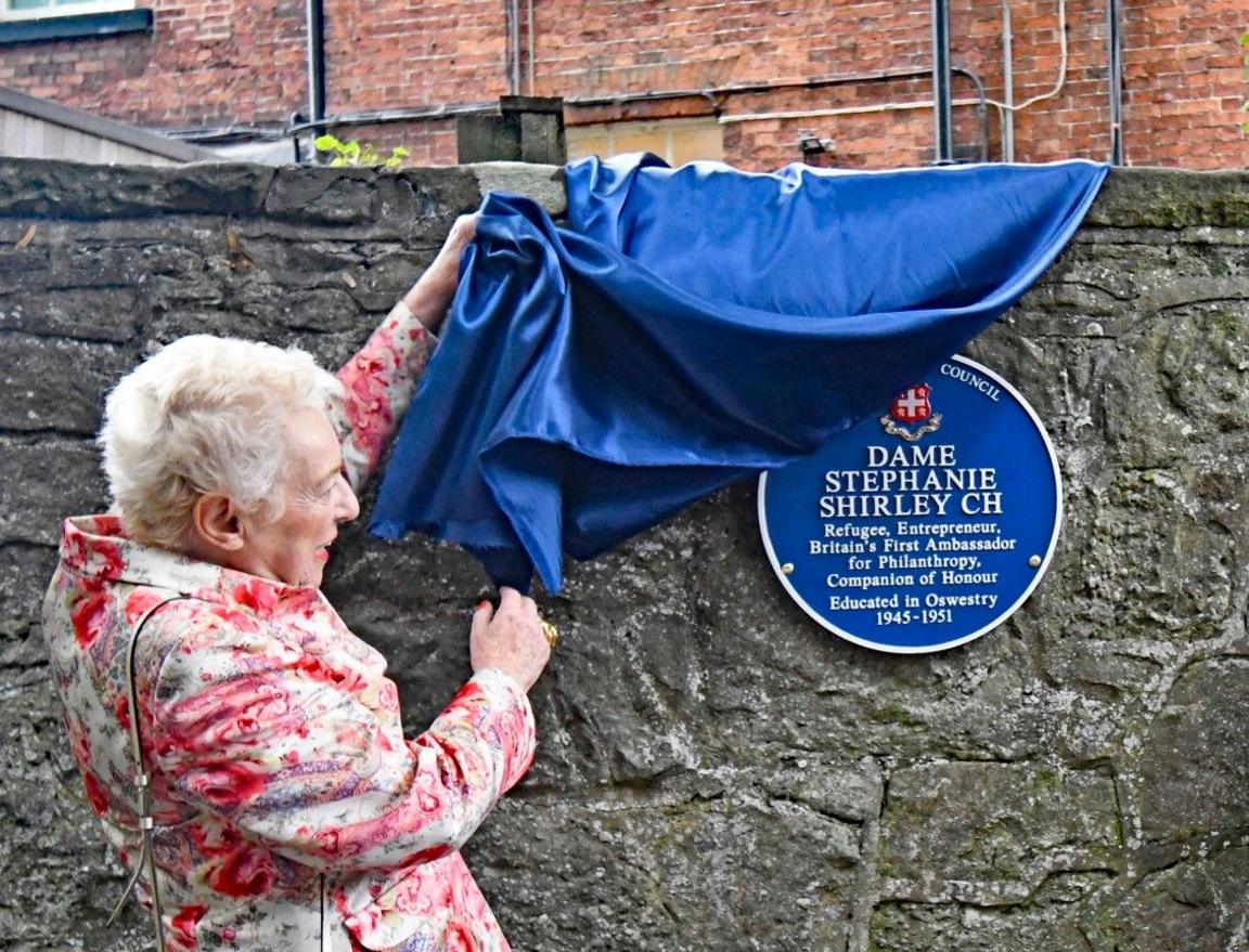 A woman unveiling a blue plaque on a wall. It reads "Dame Stephanie Shirley CH". "refugee, entrepreneur, Britain's first ambassador for philanthropy, companion of honour"