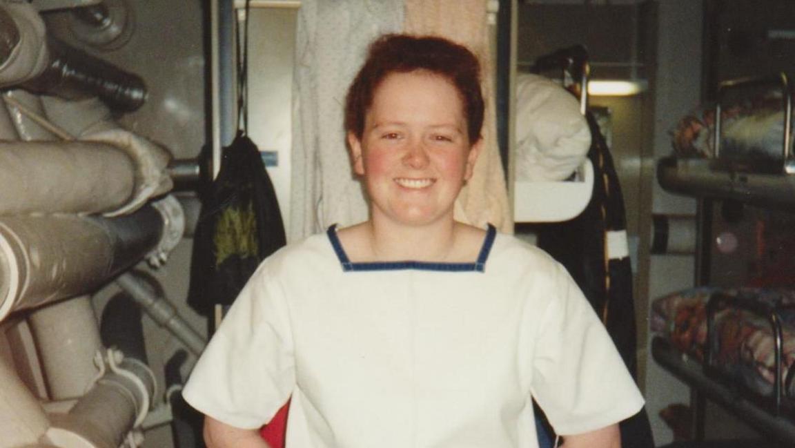 Picture taken in the early 90s of a young red-head in her Navy uniform. She is stood next to her bunk and smiling at the camera.