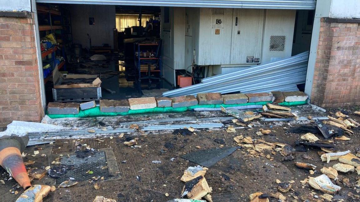 Rubble and debris are on the ground outside a brick building with an opening to the warehouse beyond with its shutters half raised.