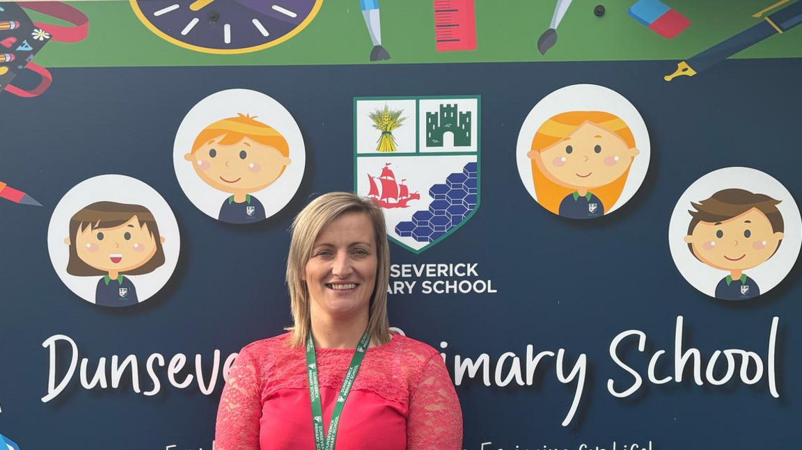 Leanne Smyth is wearing a pink top with lace sleeves and has a green lanyard around her neck. She is standing in front of the sign for Dunseverick Primary School which displays a school crest and four cartoon-style portraits of pupils: two bots and two girls.