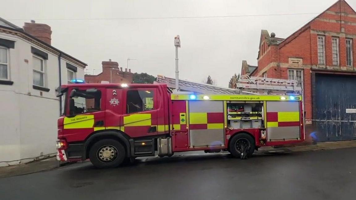 A fire engine with blue lights on. It is parked in a street with a large brick building to one side and a white house on the other.