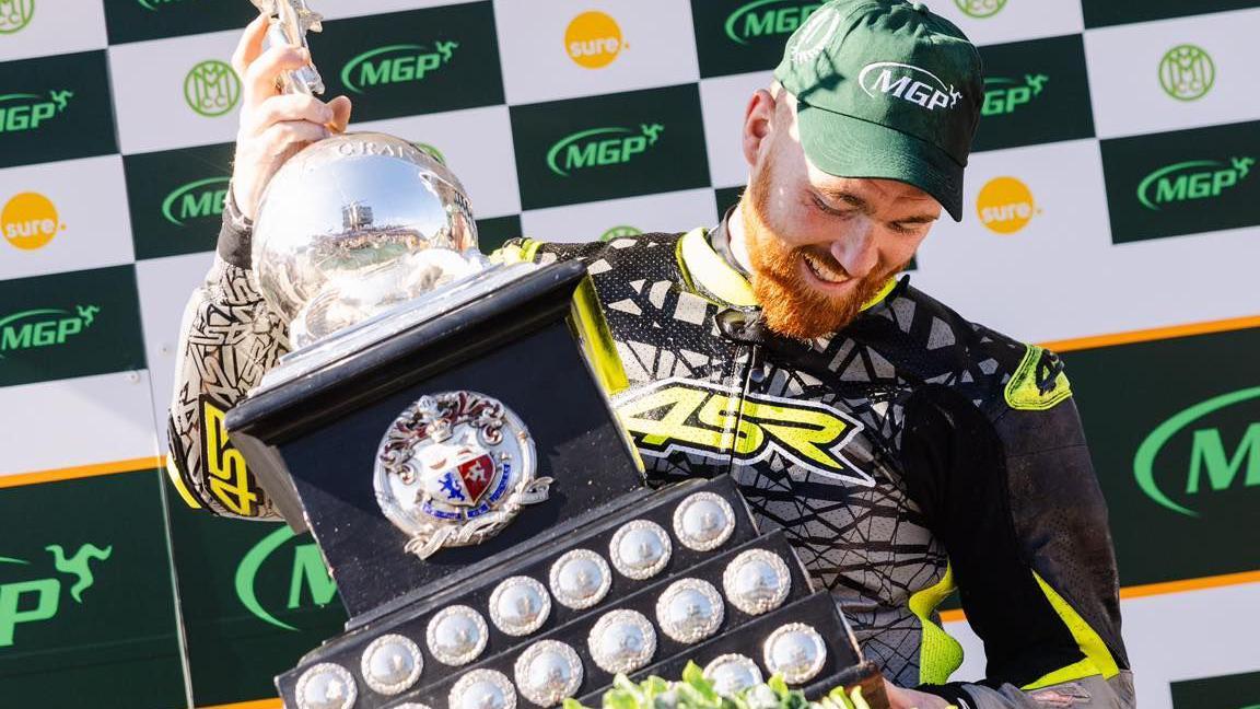 Caomhan Canny holds the Senior MGP trophy. he's wearing black leathers with yellow trim and a green MGP cap.
