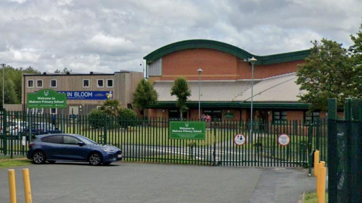 Malvern Primary School is a redbrick building set behind green metal fencing. A green sign with white writing reads Welcome to Malvern Primary School.