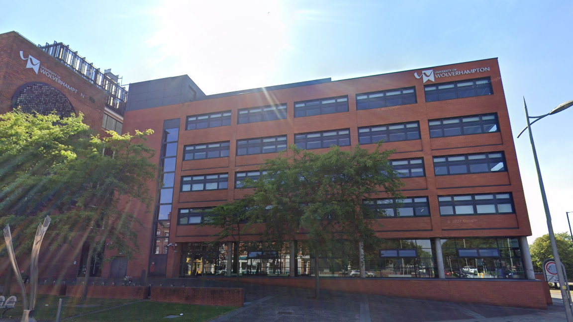 The front of a university of Wolverhampton building. It is red brick and has rows of windows. There are green trees outside and the sun is peeking over the roof of the building. A silver sign on the building says "university of Wolverhampton"