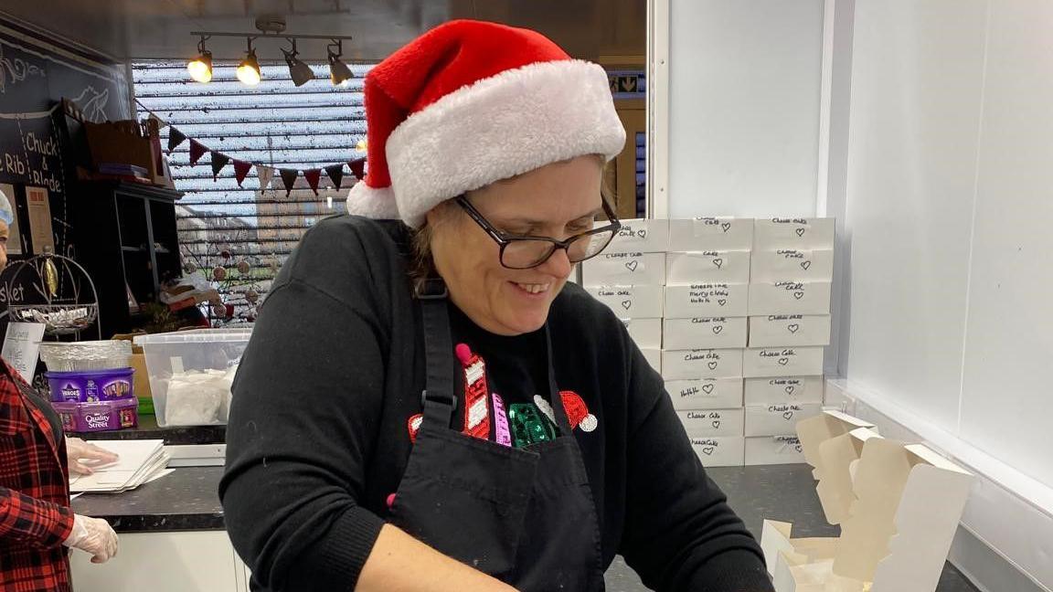 A woman stands with a Christmas hat on - she is cutting up some puddings and you can see piles of boxes behind her 