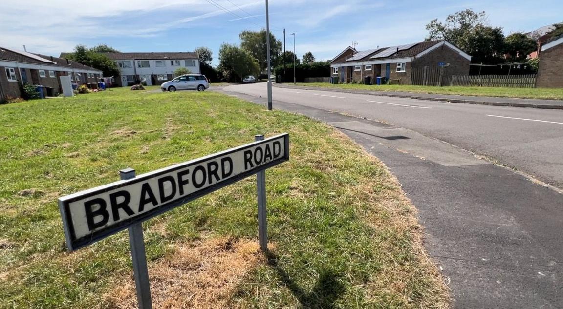 A black and white street sign on a verge at end of Bradford Road, with houses in the background.