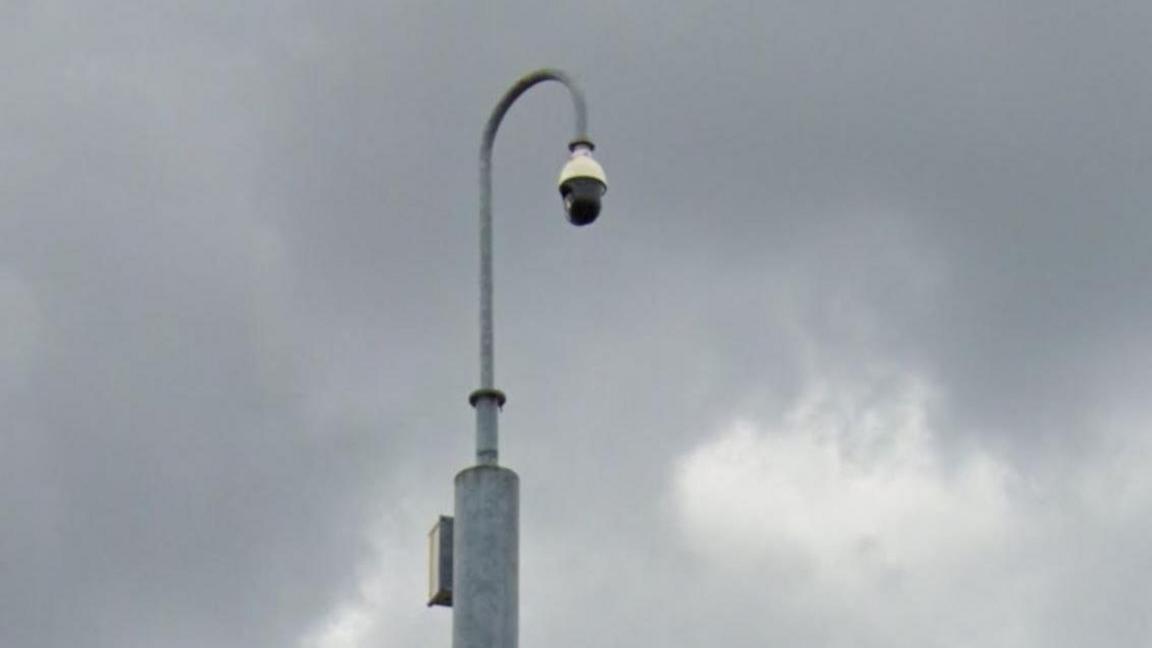 Close up of a black domed camera hanging from a hooked grey lamppost