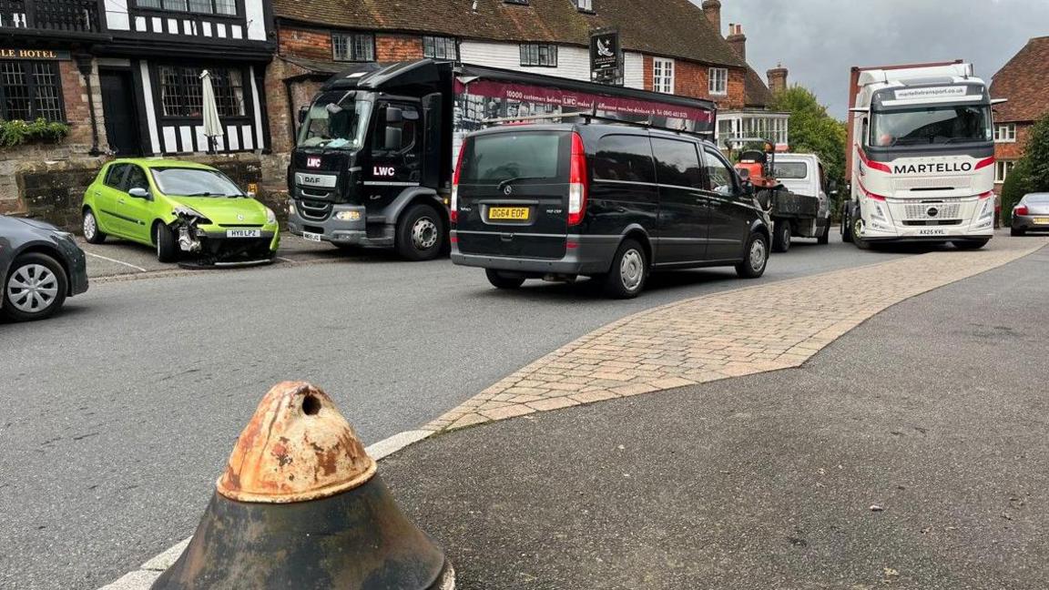 A close up of Goudhurst High Street with a large white HGV in the background beginning to make it's way up the hill towards a tight section of road. A significantly damaged green car can be seen in the background parked in front of some listed buildings.