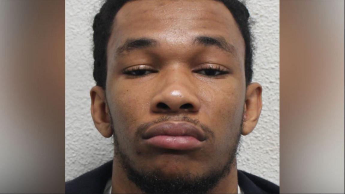 A police mugshot of Kami Carpenter shows him looking at the camera with a sullen expression. He is wearing a white T-shirt and a black layer.