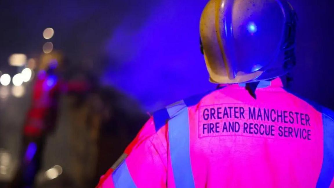 The back of a Greater Manchester Fire and Rescue Service firefighter wearing a gold helmet and a pink and blue uniform with the background blurred out