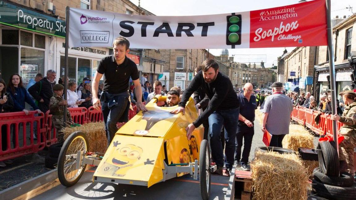 Two men push a yellow Minion-themed cart at the start line with the driver's head just visible out of a hole in the top.