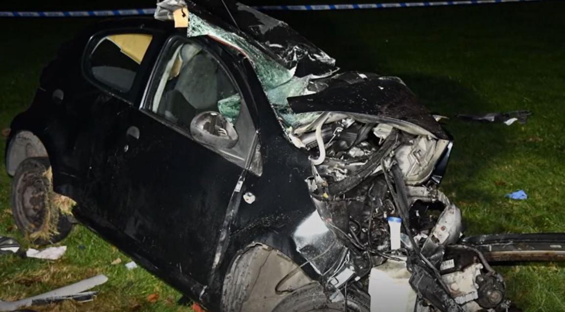 Wreckage of the dark-coloured Citroen C1 in the aftermath of the crash. The wheels are misaligned, with the roof and front of the vehicle crumpled by the impact