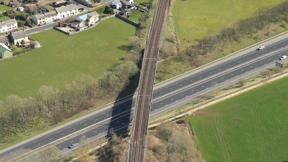 An aerial view of Clifton Bridge going over the M6. It is a long, two-track railway bridge, with residential houses to the left. 