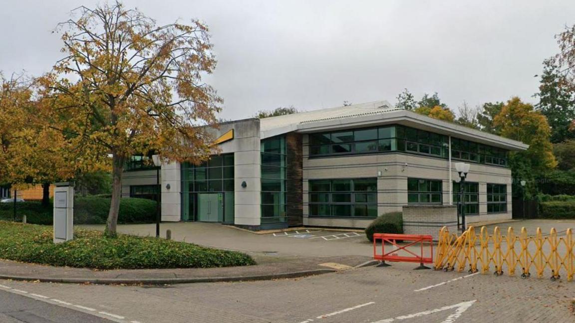 An office block is grey with large windows. It is two storeys high and there is a small parking area at the front. There is a tree and some foliage near the road, and barriers preventing access to another road