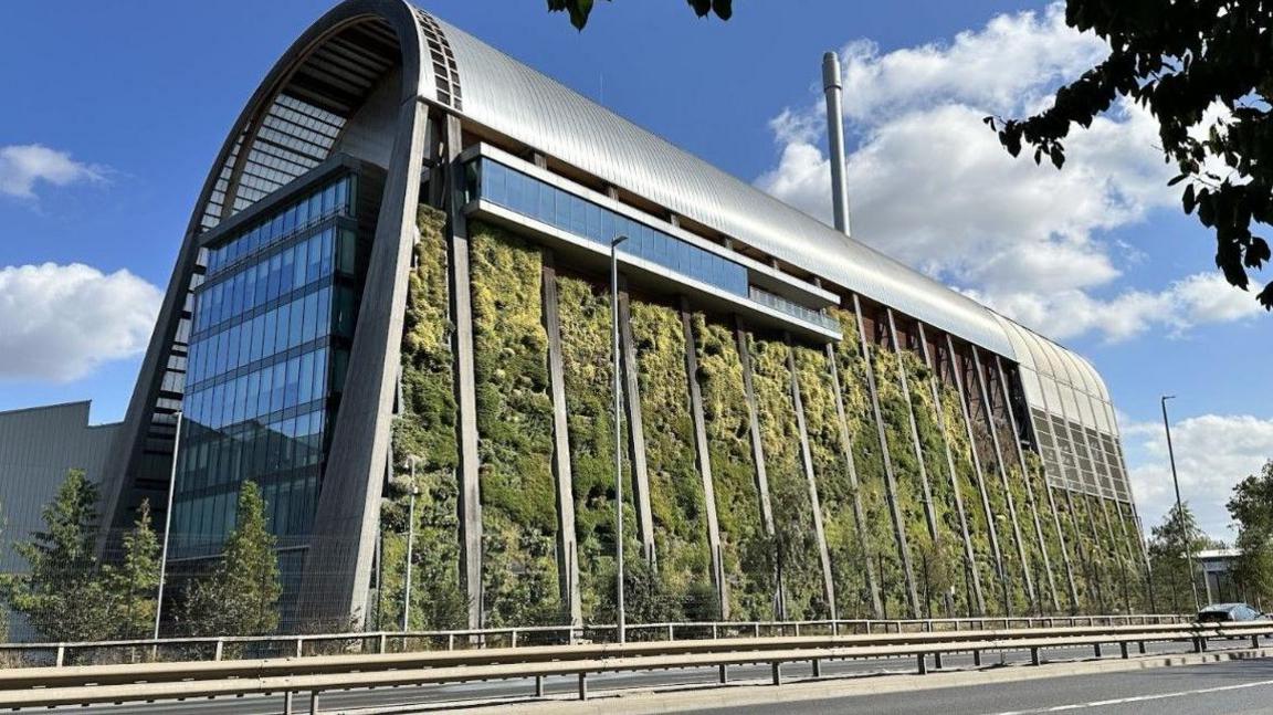 A large, modern building with a curved metallic roof. The front facade is covered with a living wall of plants. In the foreground there is a road with a metal barrier running through the centre of it.