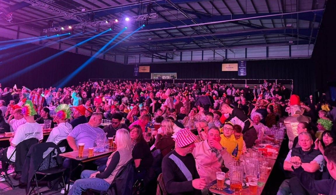 People are sitting at tables, which have beer glasses on them. The bottom half of the image is packed with people, some in hats and fancy dress, with the ceiling above them.