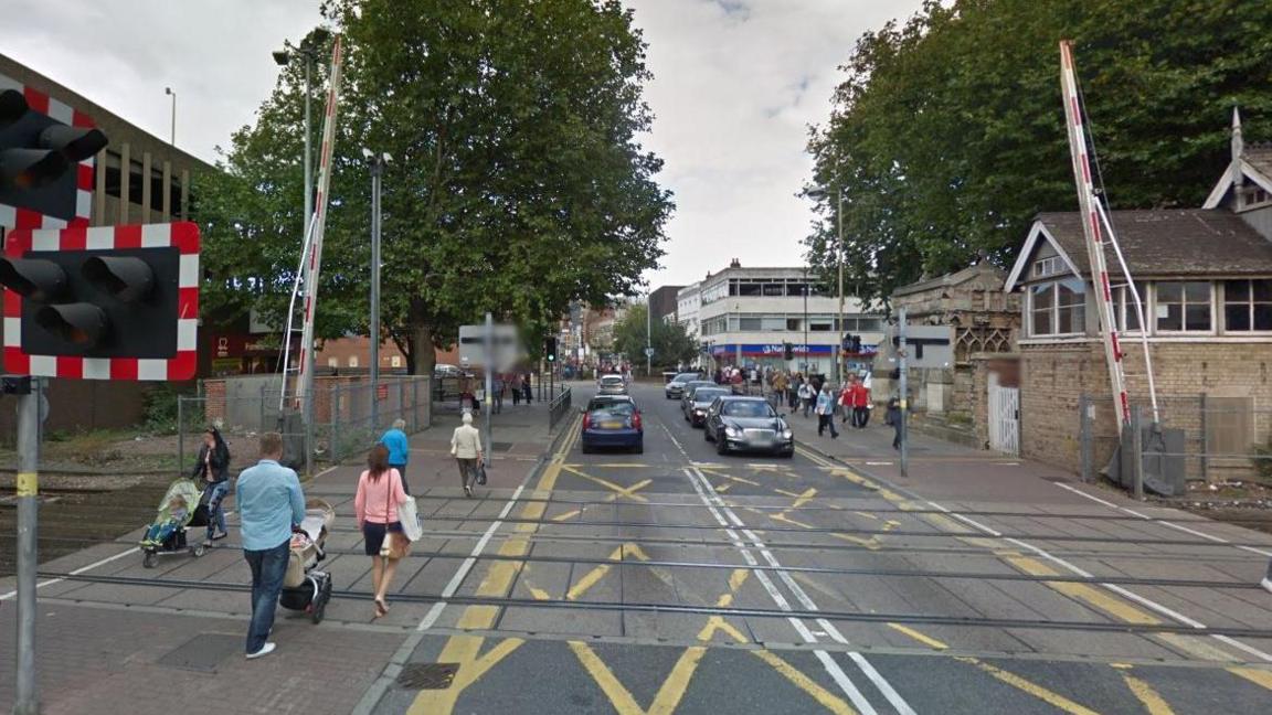 Eight pedestrians walking across a level crossing near a train station. There are yellow road markings and cars on either side of the road in the background