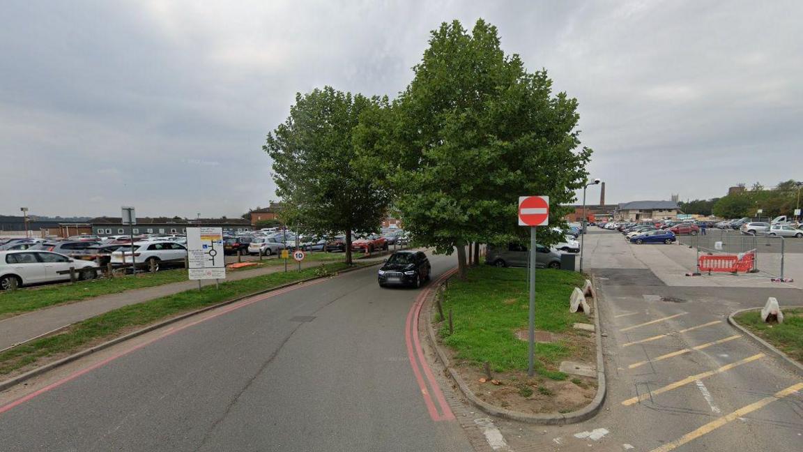 An exit road from large, busy car parks at Lincoln County Hospital. Two green trees dominate the centre of the photo and a no-entry sign stands in front of them. A black car is travelling beneath the trees as it leaves